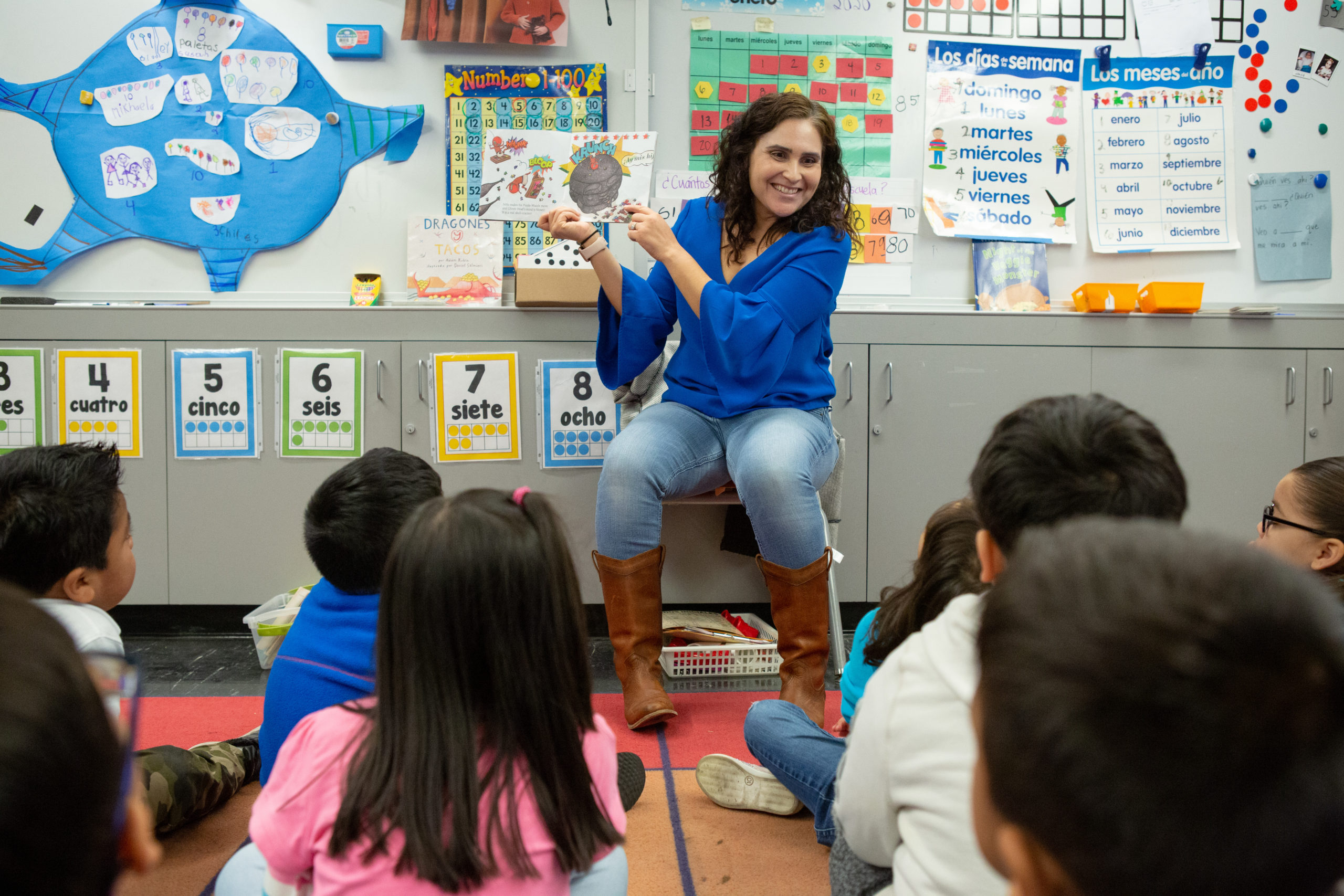 A teacher in a blue shirt reads a book to a group of young students sitting on the floor in a colorful classroom.