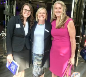 Three women smiling, standing together dressed in business attire, one in a black suit, another in a cardigan, and the third in a pink dress.