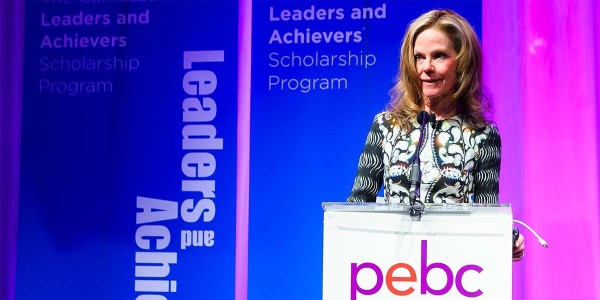 A woman speaks at a podium with "pebc" and "Leaders and Achievers Scholarship Program" banners in the background.