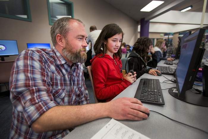 A bearded man in a plaid shirt helps a young girl in a red hoodie use a computer in a classroom setting.