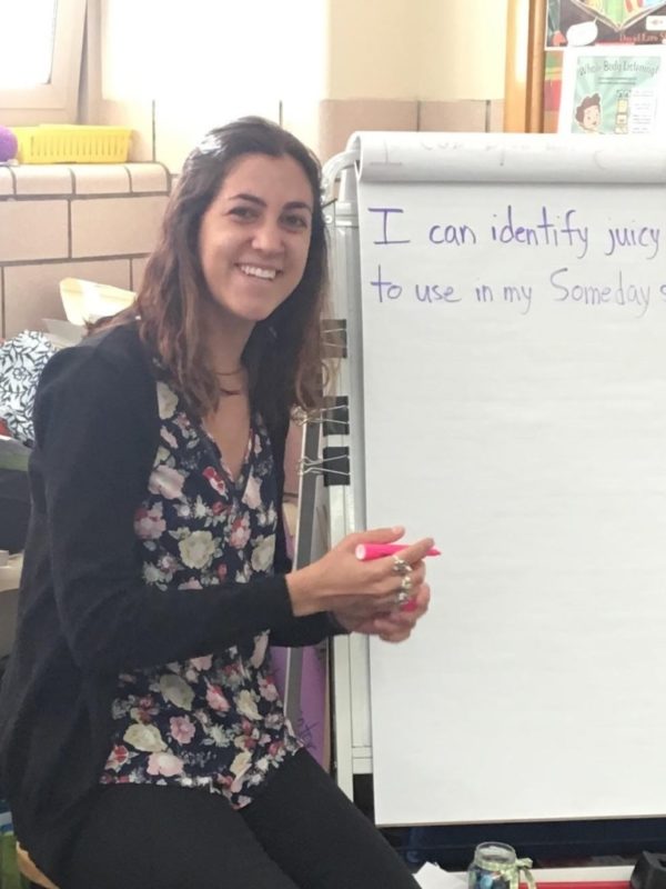 A woman with long brown hair smiles while sitting next to a whiteboard with writing on it.