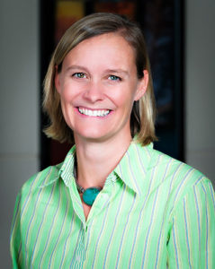A woman with short blonde hair, wearing a green striped shirt and a blue necklace, smiles at the camera.