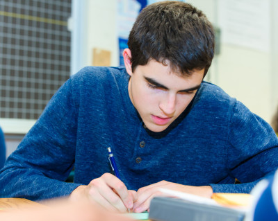 Young man in a blue shirt writing at a desk in a classroom setting.