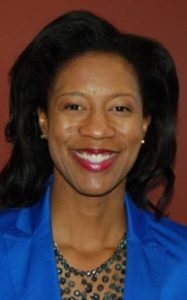 A smiling woman with long dark hair, wearing a blue blazer and a necklace, stands against a maroon background.