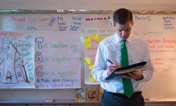A teacher in a green tie writes on a clipboard in front of a classroom whiteboard with various educational notes.