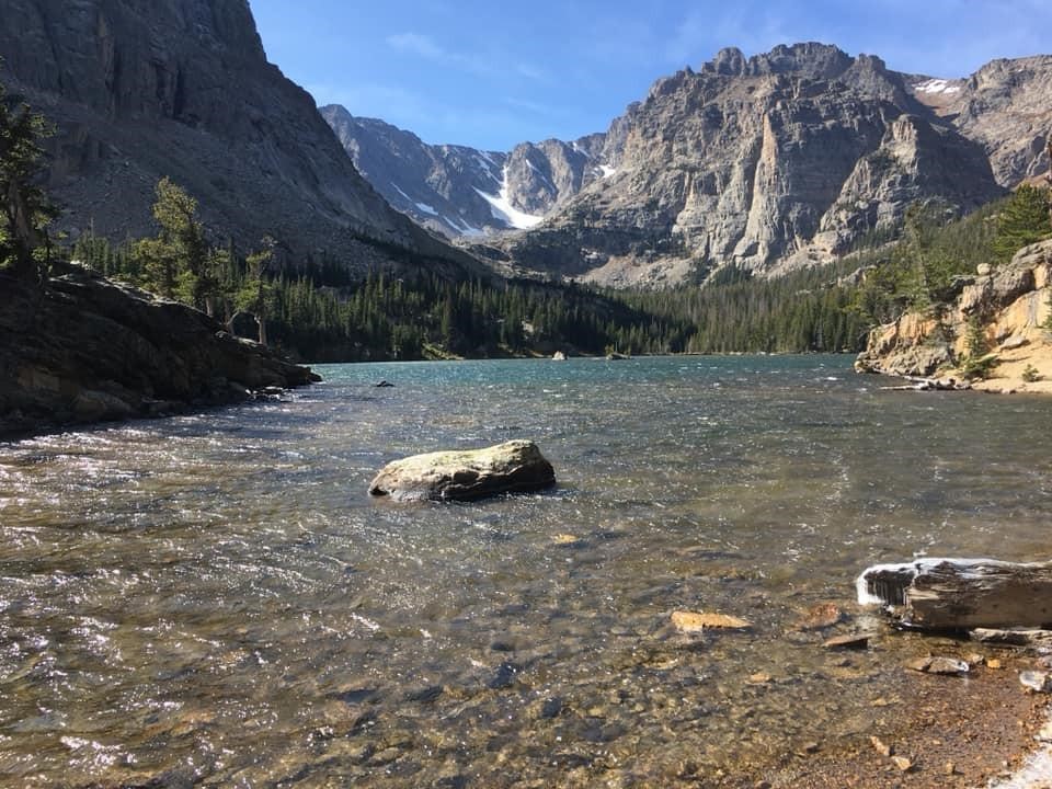 Serene lake with clear water, surrounded by rocky mountains and pine trees under a clear blue sky.