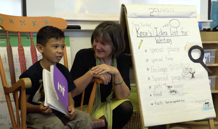 A young boy shares a book with a woman in a classroom. A flip chart next to them lists writing ideas.