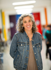 A person with gray curly hair wearing a denim jacket smiles while standing in a brightly lit corridor.