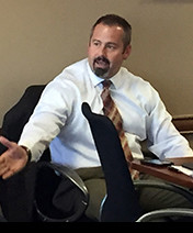 A man in a white shirt and tie, seated at a conference table, gesturing with his right hand during a discussion.