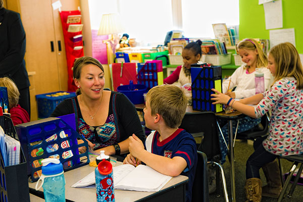 A teacher interacts with a student at a table in a brightly colored classroom; other students are engaged in activities.