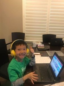 A child wearing yellow headphones gives a thumbs up while using a laptop at a table with papers and other items.