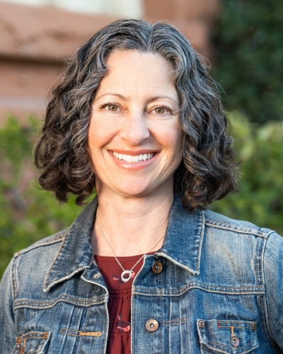 Smiling woman with curly, shoulder-length hair wearing a denim jacket and necklace, standing outdoors with greenery in the background.