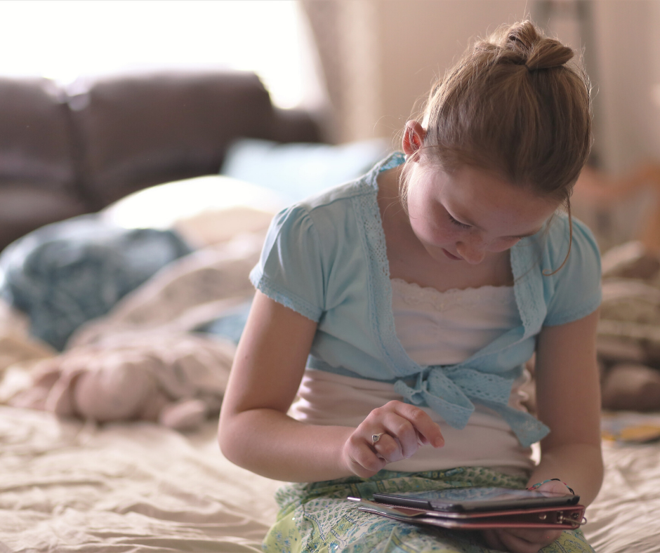 Young girl with a ponytail, wearing a blue shirt, uses a tablet while sitting on a bed in a softly lit room.