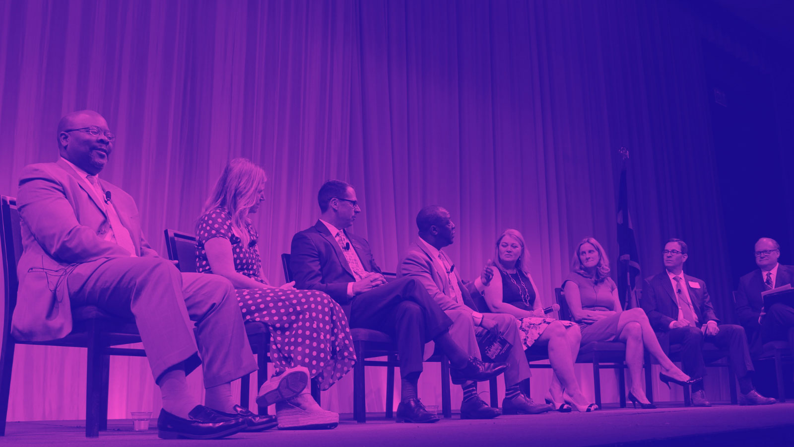 A group of eight people, dressed in formal attire, seated and engaged in a panel discussion on a stage with a curtain backdrop.