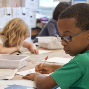Children are seated at a classroom table, focused on writing and drawing activities.