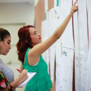 Three women stand and examine papers on a bulletin board; one points while another takes notes.