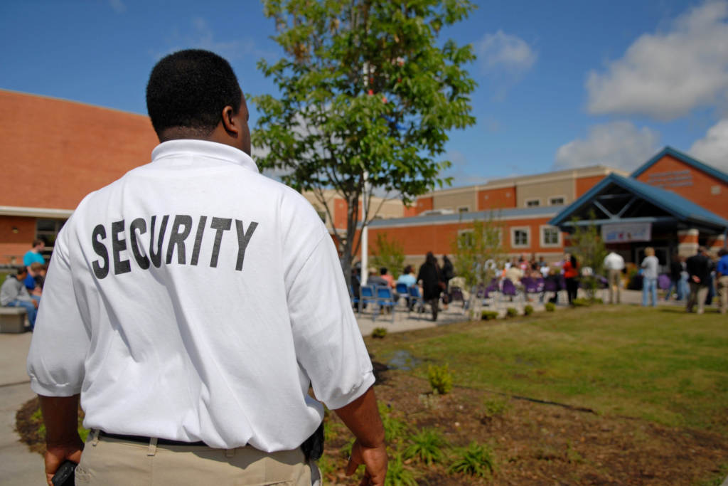 A security guard in a white shirt monitors a school event with a crowd gathered near a building on a sunny day.