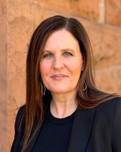 A woman with straight brown hair and a black top, standing in front of a brick wall.