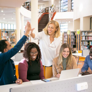 A diverse group of people high-fiving and smiling while working together in a library with laptops on the table.