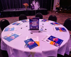 A round table with event pamphlets and a centerpiece with QR codes, set in a dimly lit room with a person seated in the background.