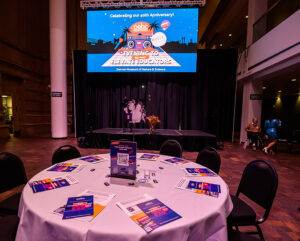 A round table with pamphlets and a QR code, set in a venue with a screen displaying an educational event announcement.