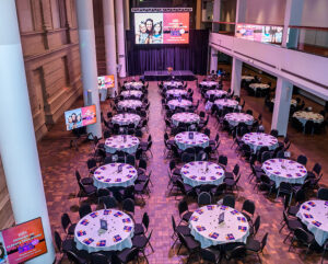 A spacious event hall set with round tables and chairs, with screens and a stage displaying vibrant signage.