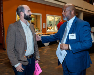 Two men in conversation at an indoor event. One holding a pink paper, and the other holding a paper and wearing a name tag.