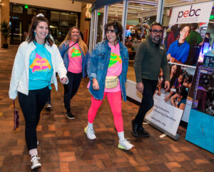 Four people in colorful outfits walking indoors, smiling and talking, with a PEBC sign visible in the background.