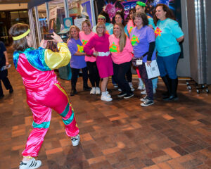 A person in a colorful tracksuit takes a photo of a group of smiling people wearing brightly colored 80s-style clothing.