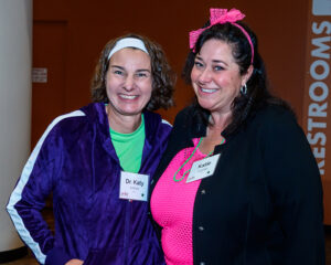 Two women smiling, wearing name tags, colorful clothing, and jewelry, standing indoors with a "Restrooms" sign in the background.