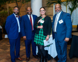 Four professionally dressed people standing indoors, smiling at the camera, with name tags and a conference setting around them.