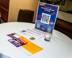 A table with pamphlets, a lit candle, and a sign with QR codes for Paddle Raise Checkout in a clean, indoor setting.