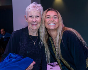 An elderly woman and a young woman with braided hair smile together, both dressed in dark clothing.