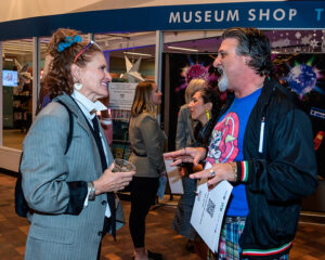 Two people engaged in conversation inside a museum shop. One holds a drink, and others are seen in the background.