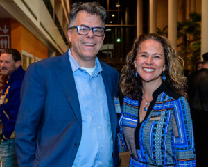 Two people, one in a blue suit and glasses and the other in a patterned blue dress, smiling at an indoor event.