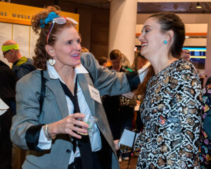 Two women are smiling and engaging in conversation at an indoor event, with other attendees in the background.