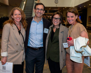 Four people smiling and posing together at an indoor event. They are holding name tags and papers.