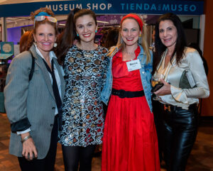 Four women are posing and smiling in front of a museum shop sign, wearing chic and varied outfits.
