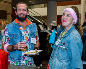 Two smiling people at an indoor event, both wearing denim jackets and 80s-themed clothing, holding drinks and plates of food.