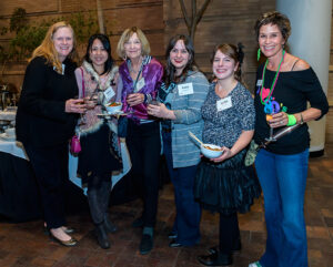 Six women, standing and smiling, holding drinks and plates of food at a social event.