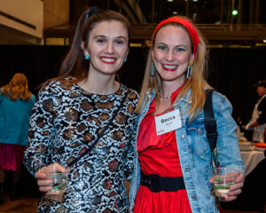 Two women smiling at an event, one wearing a patterned dress, the other in a red dress with a denim jacket and name tag.