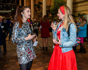 Two women conversing at an event; one in a patterned dress, and the other in a red dress and denim jacket, holding drinks.
