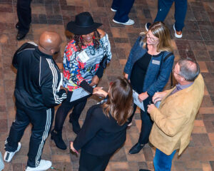 A group of people are standing together and conversing at an event, holding papers and wearing name tags.