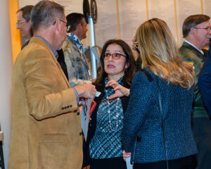 Three people are engaged in conversation at an indoor event, with others interacting in the background.