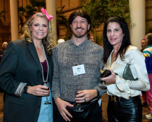Three people smiling and holding drinks at an indoor event, with name tags visible and greenery in the background.