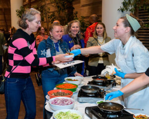 People serving food at a buffet, including a chef and guests in line with plates, in a lively, indoor setting.