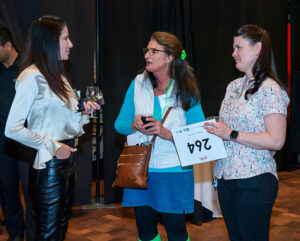 Three women at an event, one holding a glass, another with a phone and a badge, and a third with a numbered sheet.