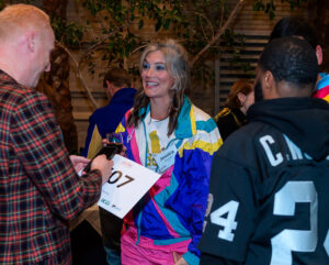 A woman in a colorful jacket, wearing a race bib, talking and smiling with a man in a plaid shirt at an event.