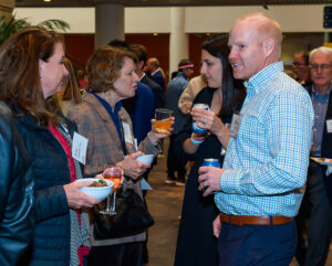 People at a social event, chatting while holding drinks and bowls of food in a venue with a casual atmosphere.