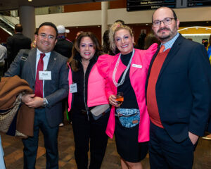 Four people posing together, dressed in business and festive attire, at an indoor event carrying name badges.
